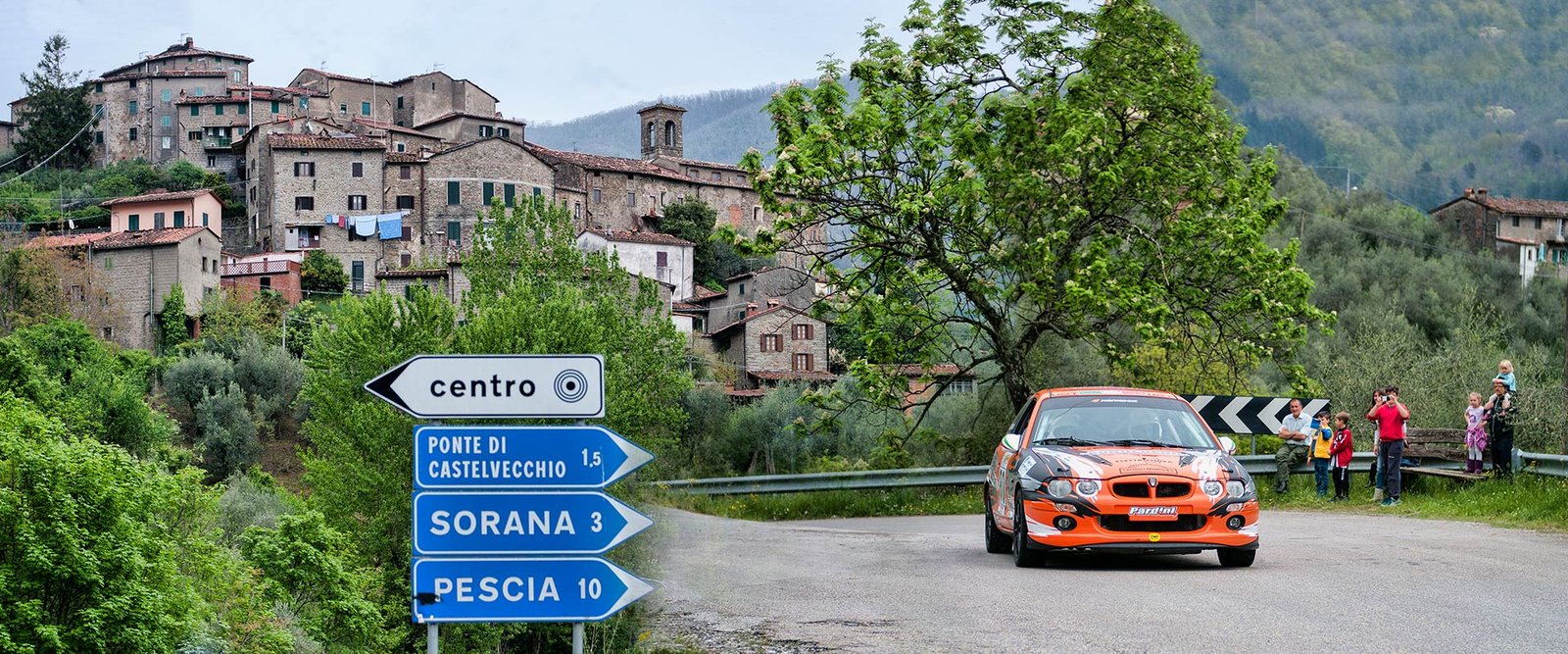 An orange rally car passing a signpost below Castelvecchio