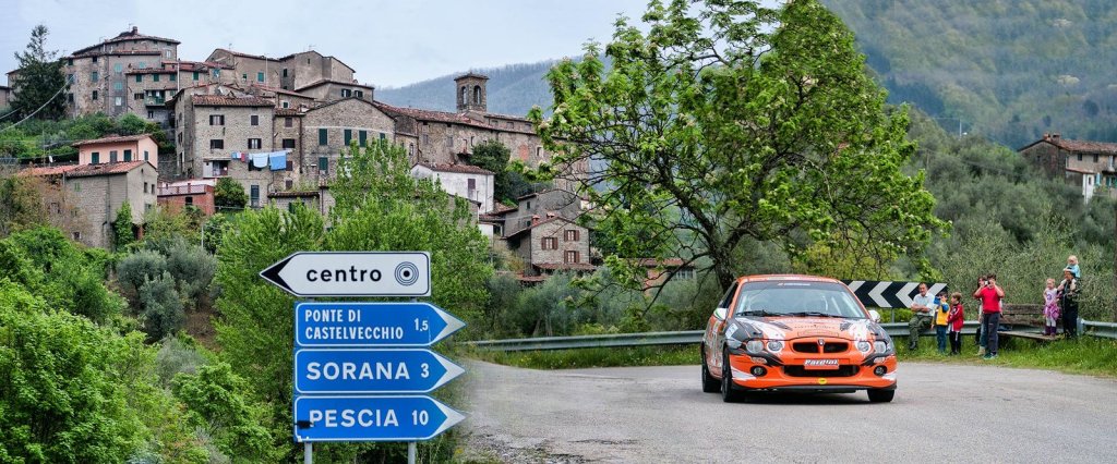 An orange rally car passing a signpost below Castelvecchio