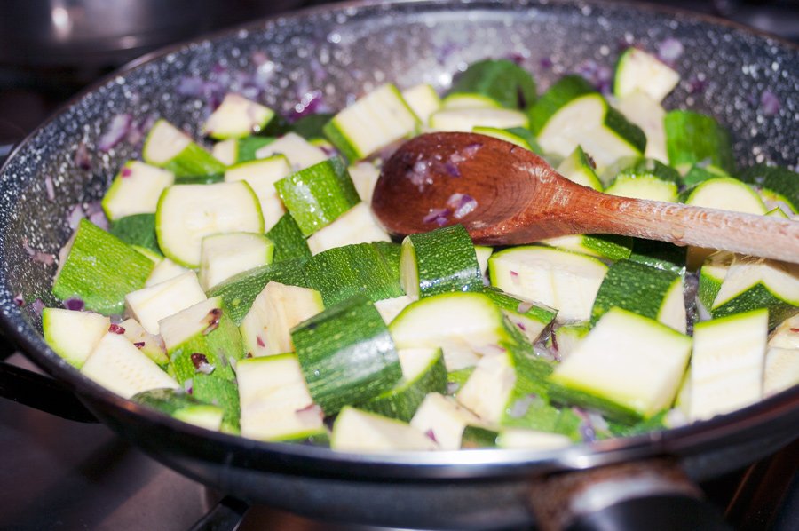 Chopped courgettes frying in a big pan with a wooden spoon