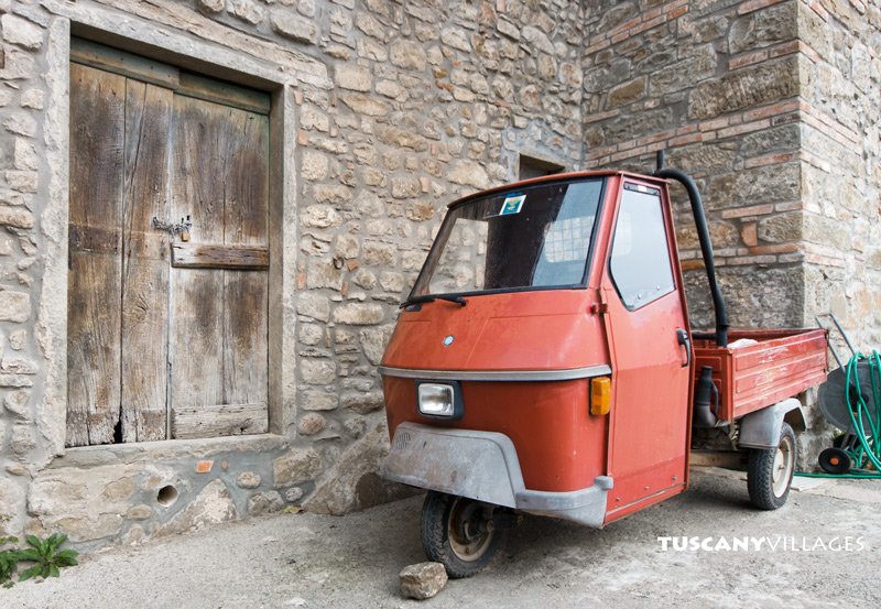 A small orange three-wheeled Ape van parked by an old wooden door in a stone wall