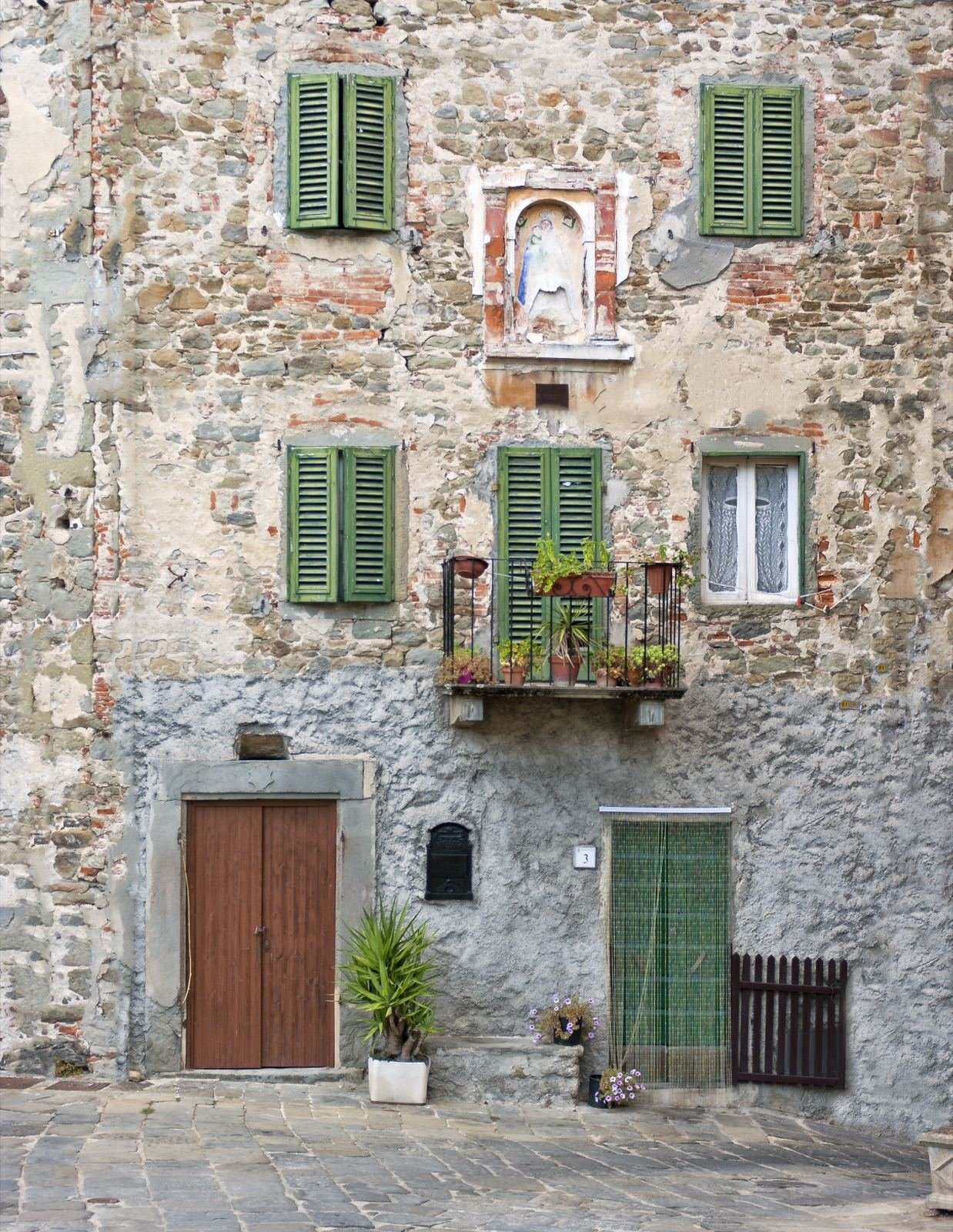 An old stone house in Pietrabuona with green shutters, a balcony of flowers and a little painted shrine on the wall
