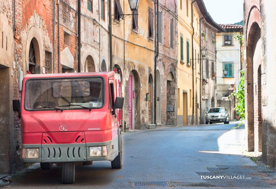 A small red van filling a narrow street of old houses in Vicopisano