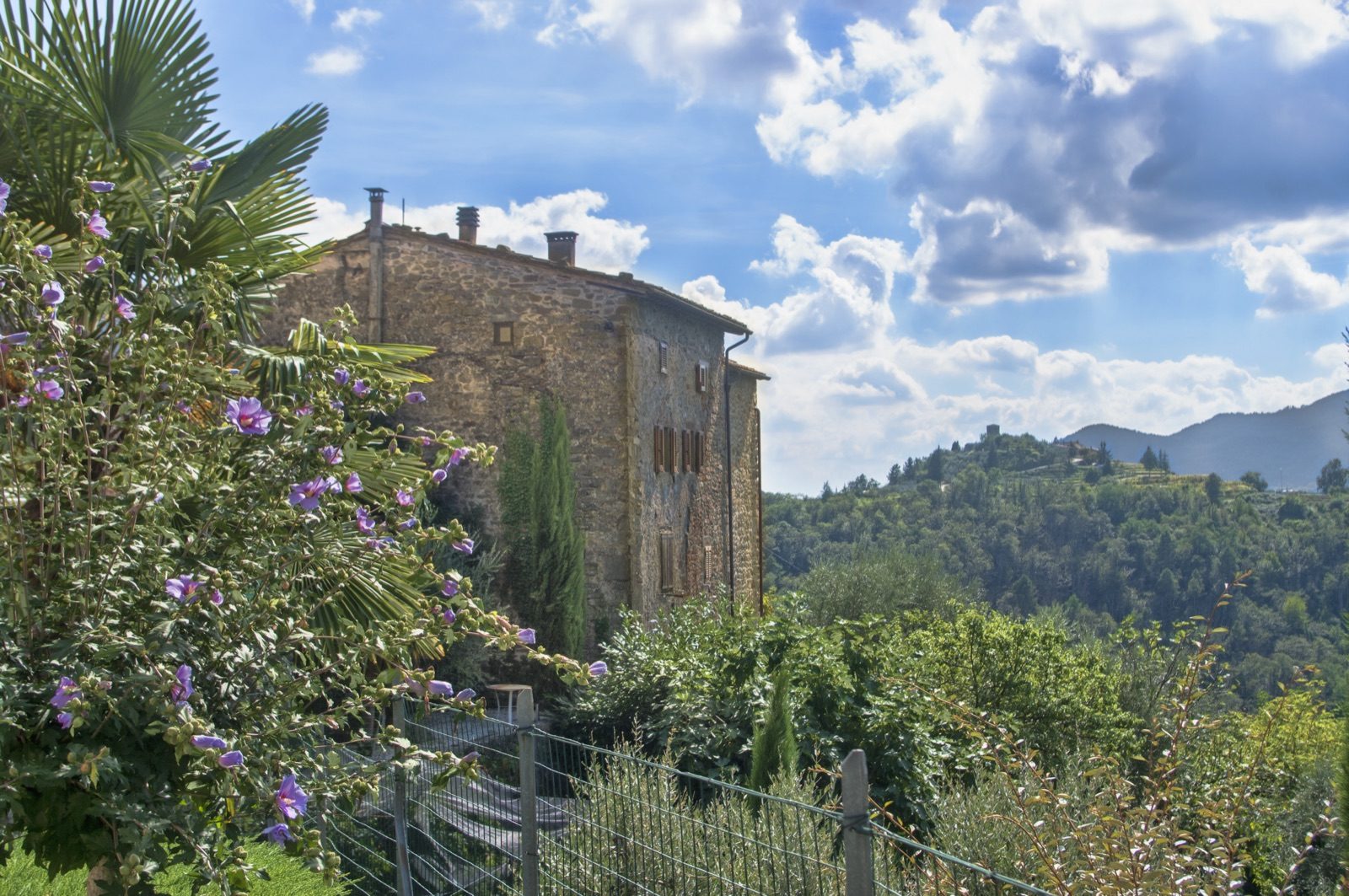 An old stone house in Sorana among purple flowers, with wooded hills and Aramo behind under a big sky